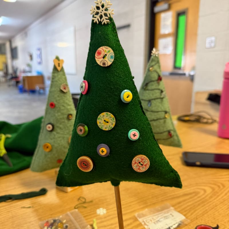 Three small, green felt Christmas trees decorated with colorful buttons stand on a wooden table in a blurred classroom setting.