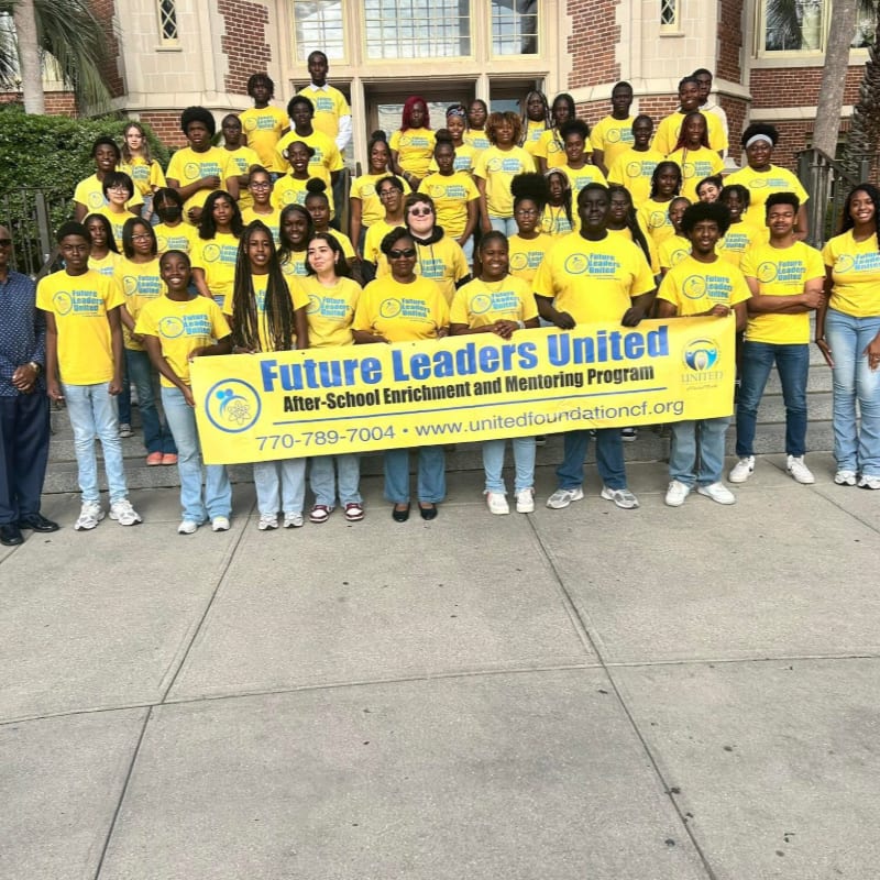 A group of students in yellow t-shirts hold a "Future Leaders United" banner on a concrete walkway in front of a brick building.