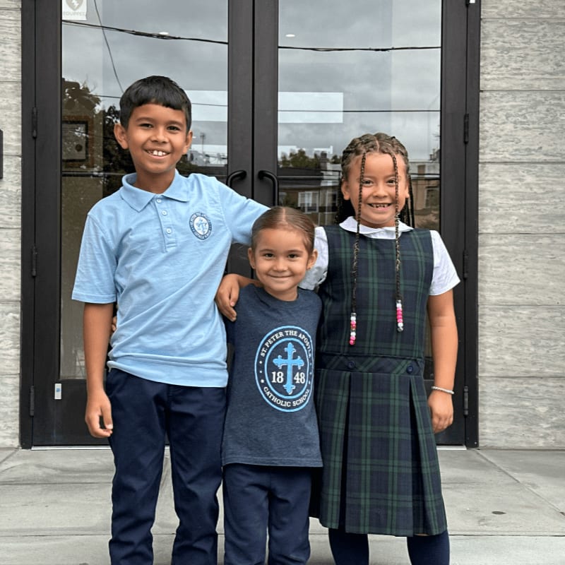 Three children in school uniforms stand smiling in the foreground in front of a set of dark glass doors.