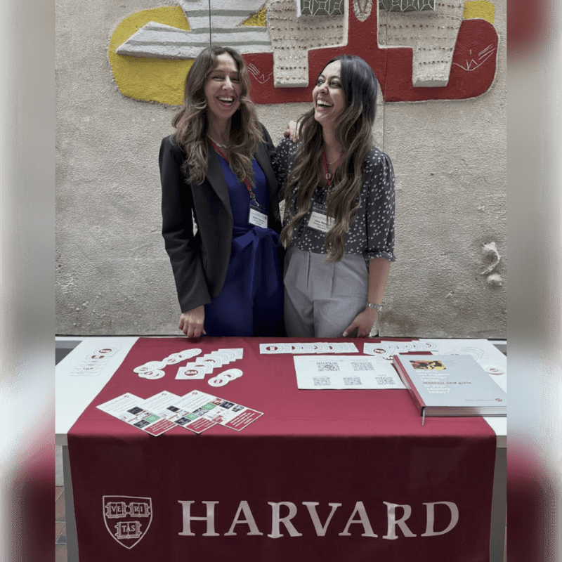 Two smiling women stand behind a red Harvard-branded table covered in brochures, set against a neutral wall with a colorful abstract mural.