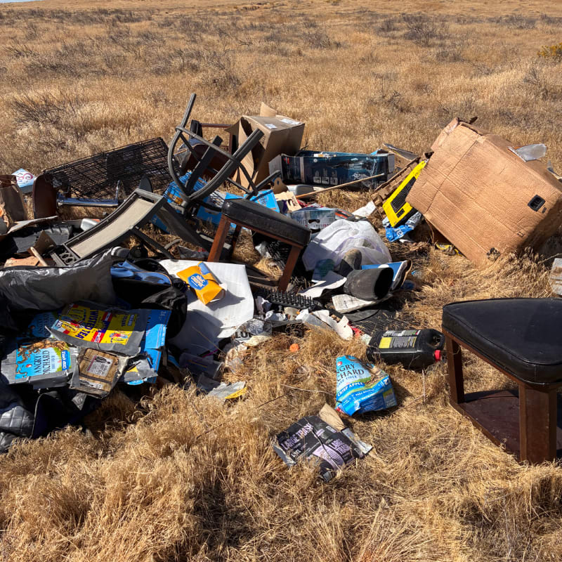A pile of discarded furniture and trash litters the foreground of a dry, grassy field.
