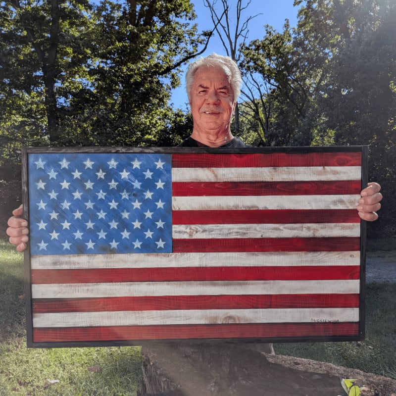 A man holds a large wooden American flag in the foreground against a background of lush green trees and a clear blue sky.