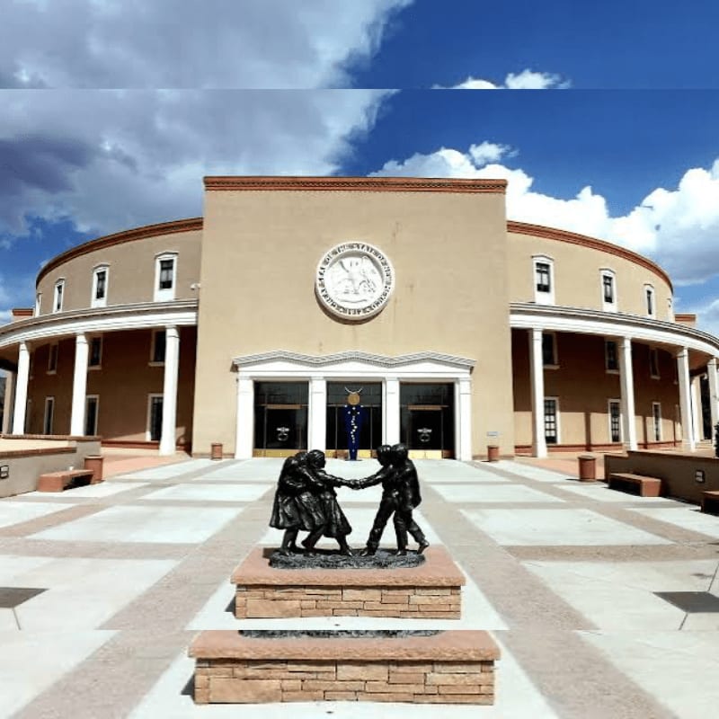 A bronze statue of two figures shaking hands stands in the foreground before a large, curved tan building under a blue cloudy sky.