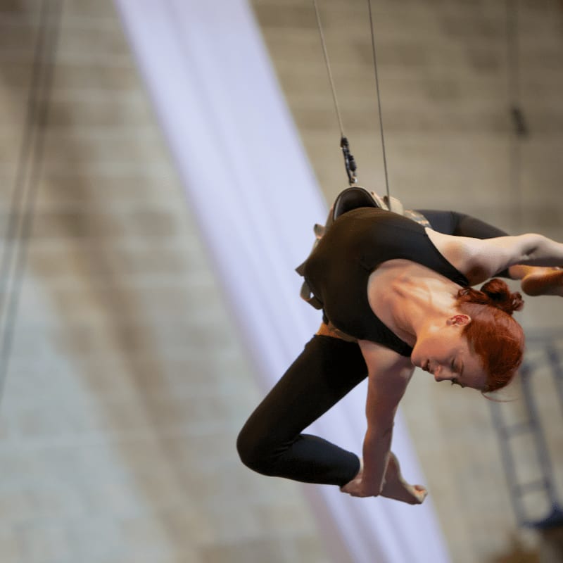 A red-haired acrobat in a black bodysuit performs a flexible pose on a trapeze against a blurred background of a white fabric drape and a concrete wall.