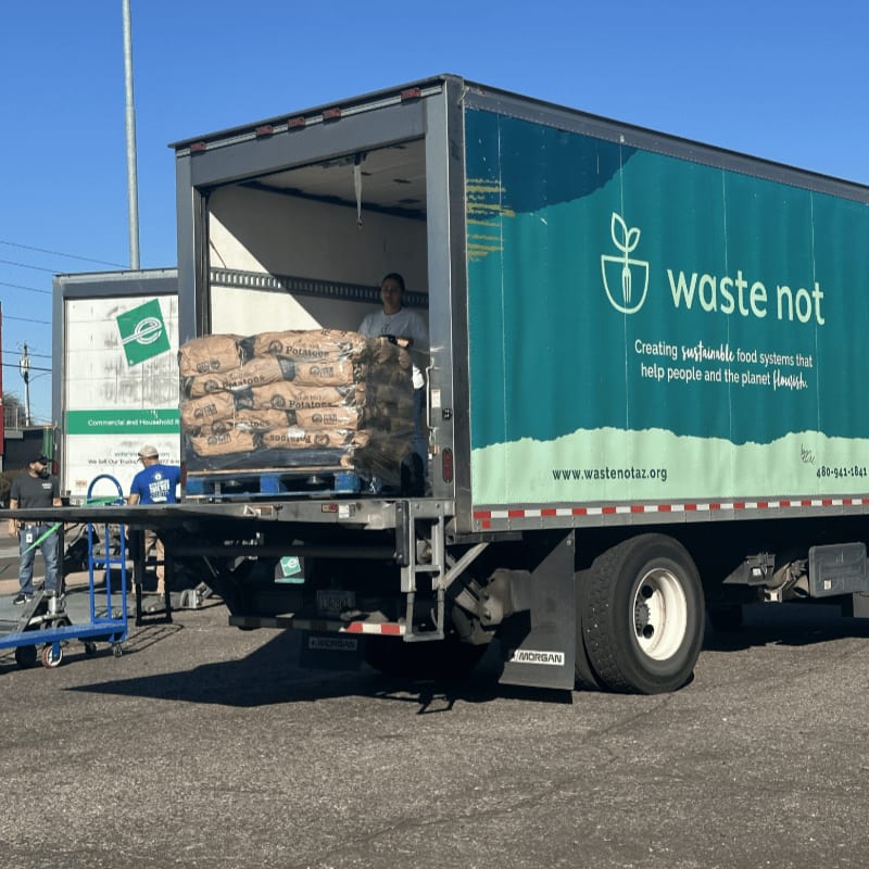 A person stands inside a "waste not" delivery truck loaded with sacks of potatoes, parked on a paved lot under a clear blue sky.