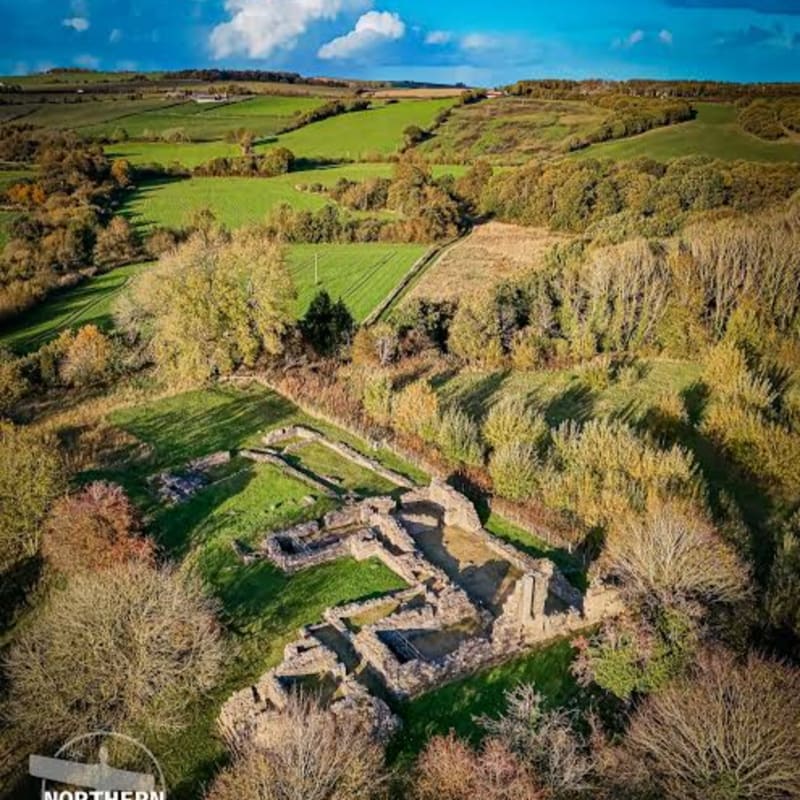 Stone ruins of an ancient structure sit in a grassy clearing, surrounded by a vast landscape of rolling green hills and autumn forests under a blue sky.