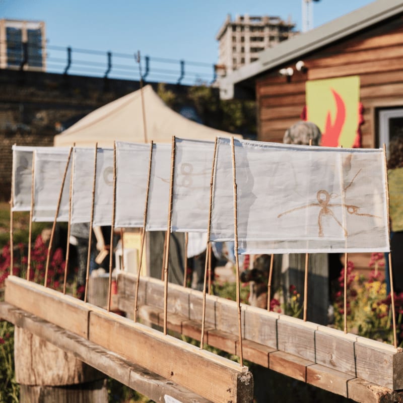 A row of white fabric banners with simple drawings stands in the foreground, with a tan tent and a wooden building set against a city skyline in the background.