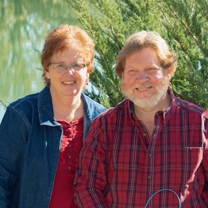 A smiling couple stands in the foreground against a background of lush green foliage.