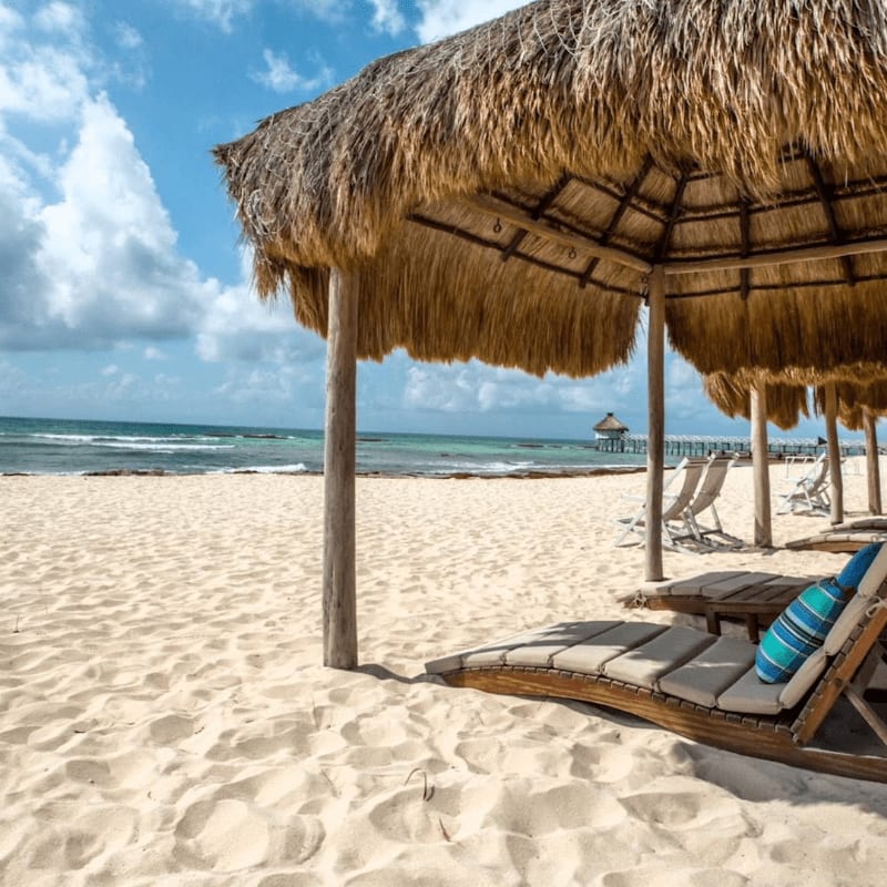 Wooden lounge chairs sit under thatched umbrellas on a sandy beach, with the turquoise ocean and a blue sky in the background.