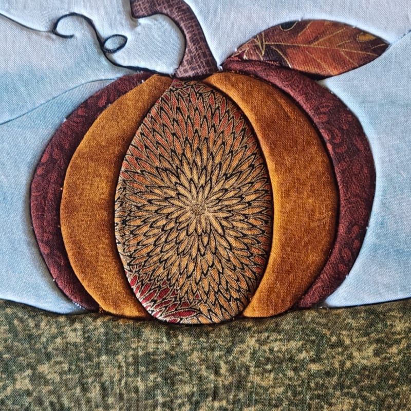 A fabric pumpkin with patterned orange and burgundy segments sits atop a textured green surface against a light blue background.