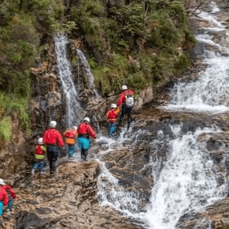 A group of people in red jackets and helmets climb across wet rocks in the foreground, with cascading waterfalls and lush green foliage in the background.