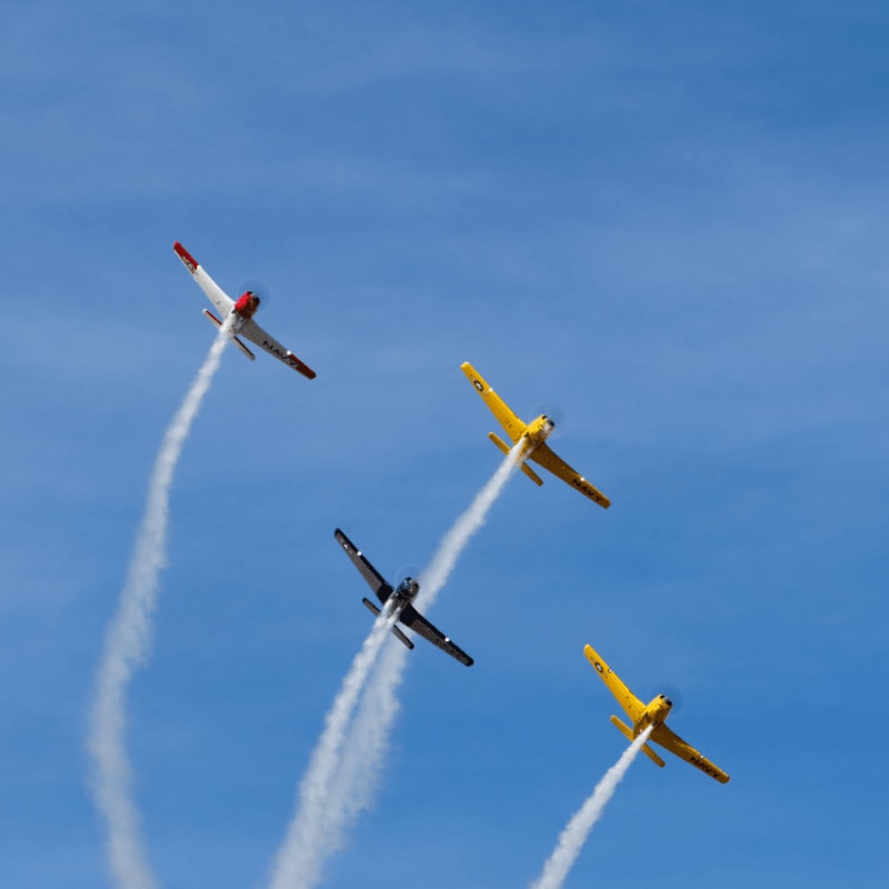 Four colorful stunt planes leave white smoke trails as they fly across a clear blue sky.
