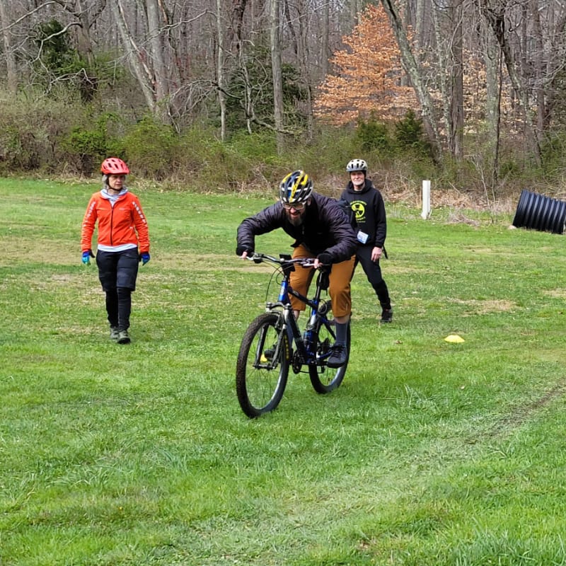 A man rides a bicycle across a grassy field while two people watch in the background against a backdrop of leafless woods.