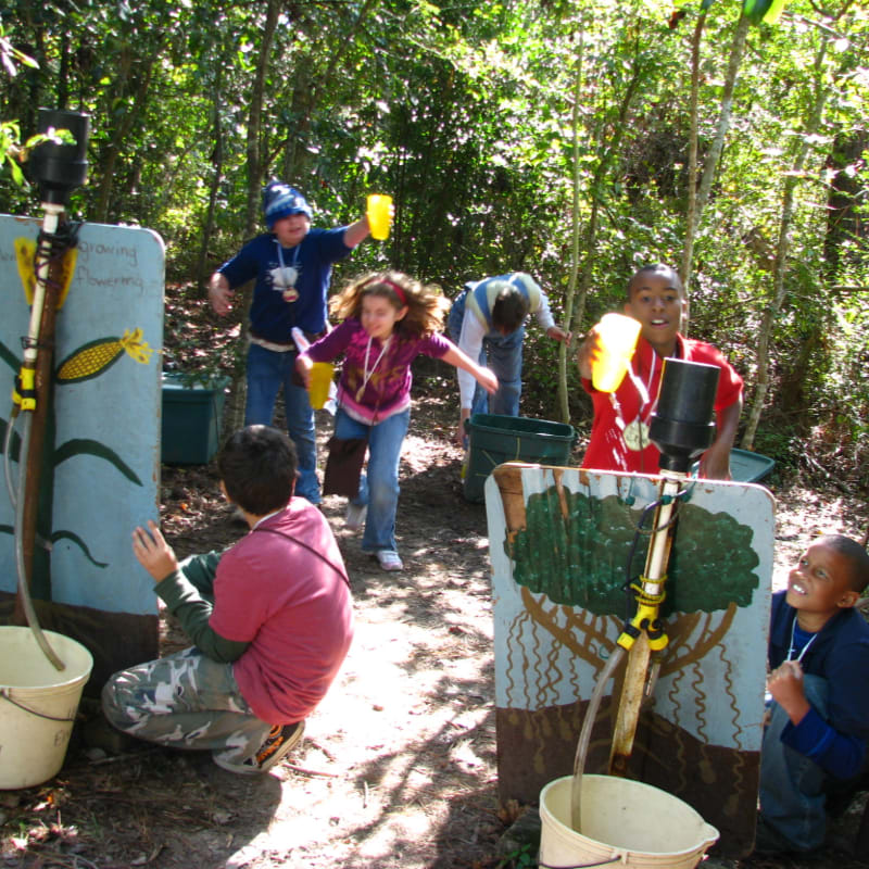 Children play a water-carrying game with buckets and cups in a wooded outdoor area.