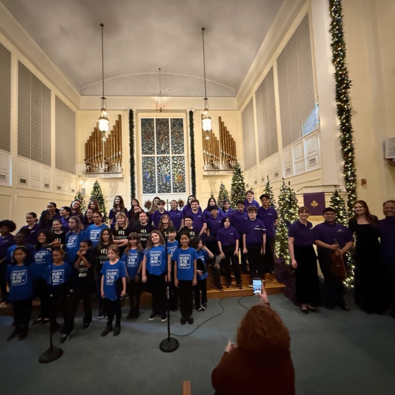 A choir of children and adults in purple shirts stands on a stage in a church, with a large pipe organ and stained glass window in the background.
