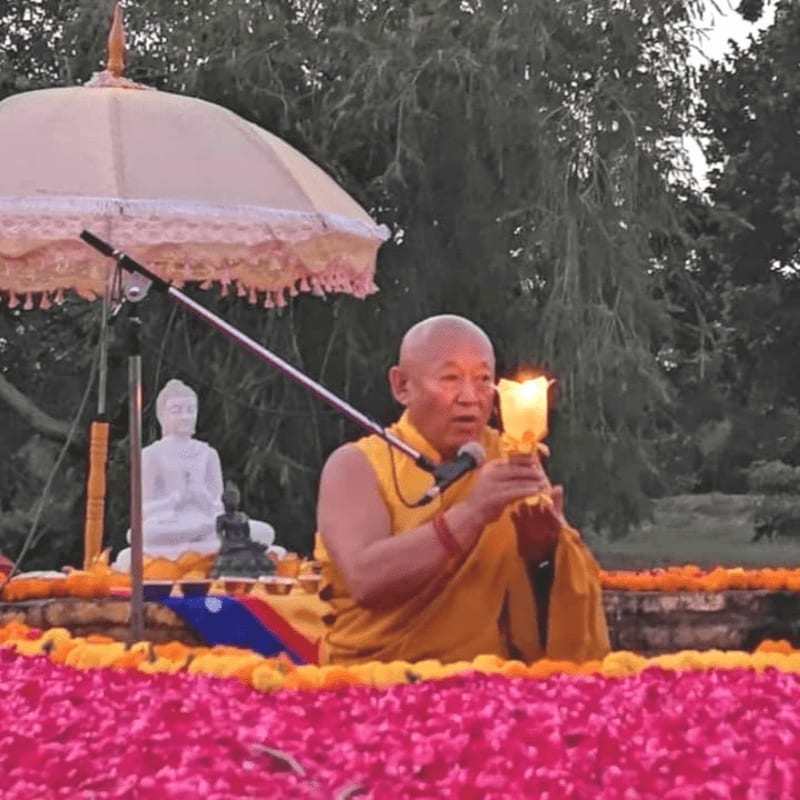 A Buddhist monk in saffron robes holds a flaming torch in the foreground, with a white Buddha statue and lush green trees in the background.