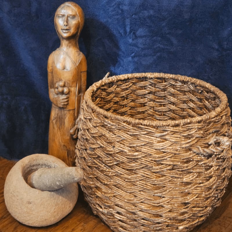 A woven basket, a stone mortar and pestle, and a wooden figurine stand against a dark blue fabric background.