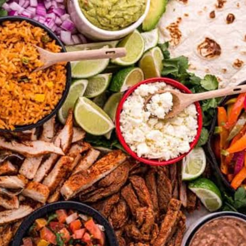 A variety of Mexican meats, rice, and toppings fill the foreground, with tortillas, guacamole, and diced onions in the background.