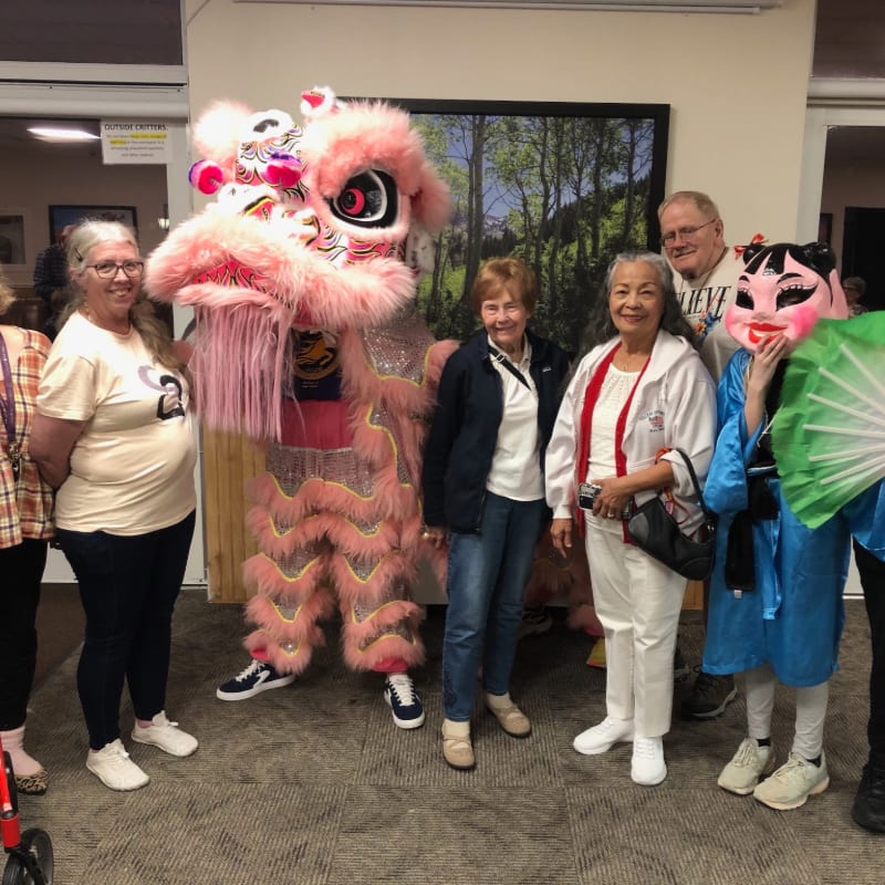 A group of people and a person in a pink lion dance costume stand together in a room with a large forest picture on the wall.