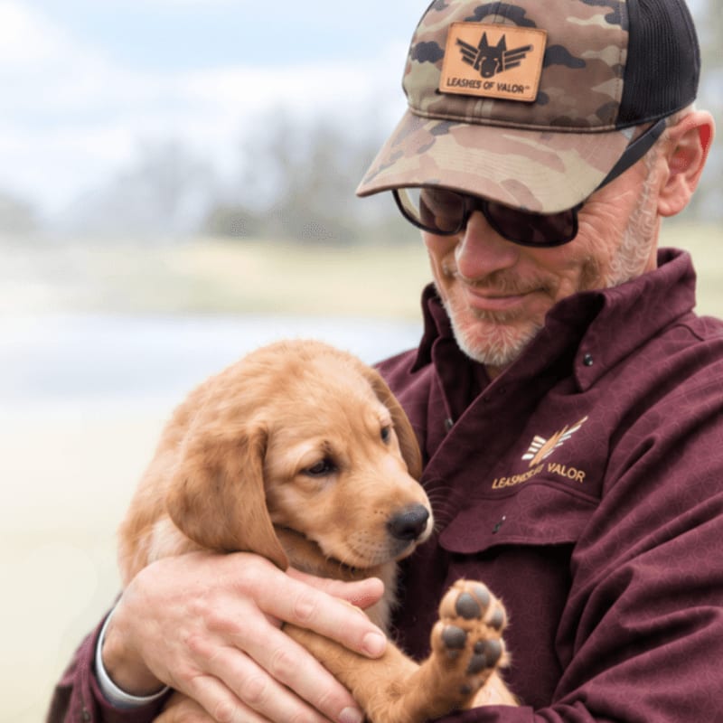 A man in a camouflage hat and maroon shirt holds a golden retriever puppy against a blurred outdoor background.
