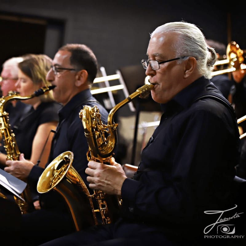 A row of musicians in black attire play saxophones in the foreground against a blurred background of other orchestra members and equipment.