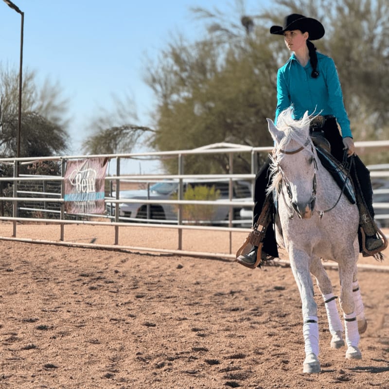 A woman in a blue shirt and cowboy hat rides a white horse across a sandy arena with a white fence and sparse trees in the background.