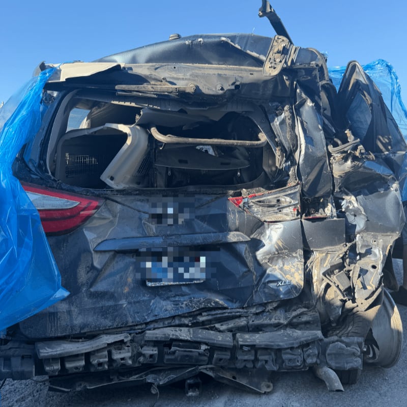 A severely wrecked dark-colored car sits in the foreground against a clear blue sky.