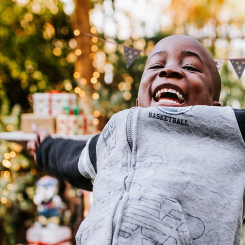 A laughing young boy in a grey zip-up hoodie stands in the foreground against a blurred background of festive lights, wrapped gifts, and greenery.