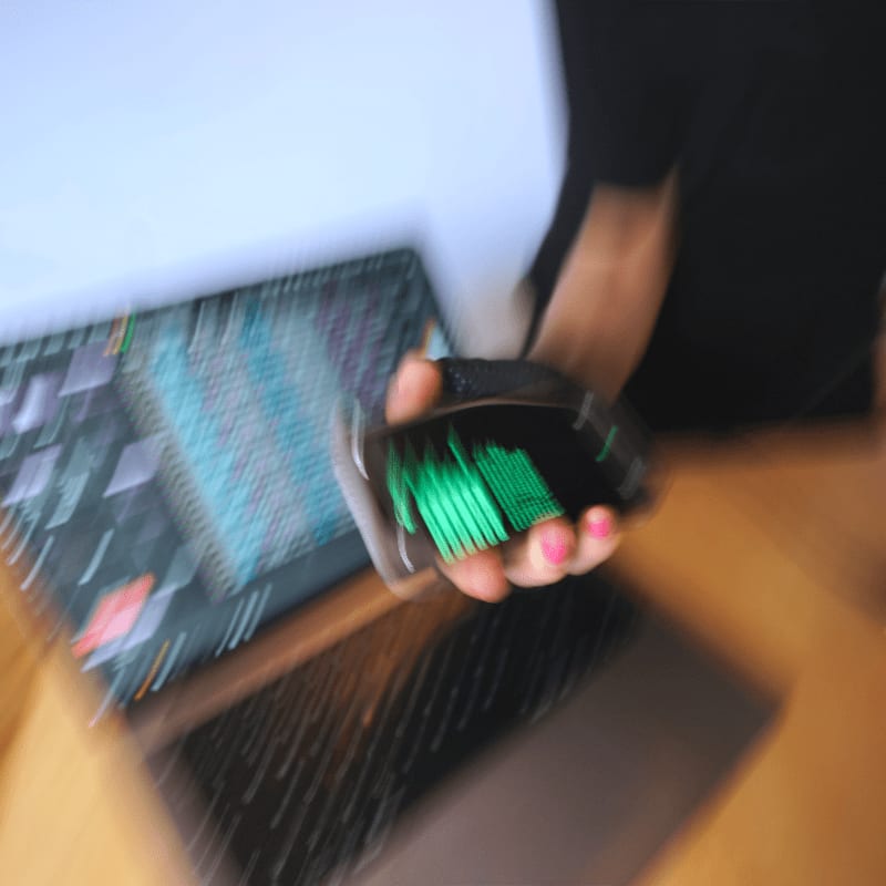 A hand holds a wallet filled with green cards in the foreground, with a blurred laptop and person in the background.