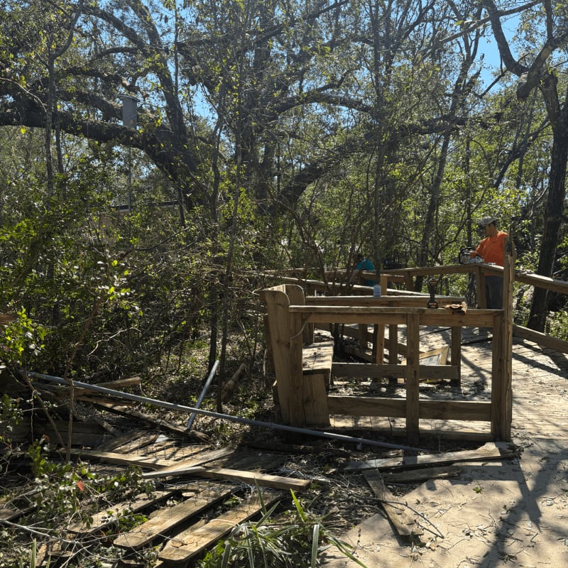 Broken wooden planks and debris litter the foreground, while a man works on a wooden railing in a wooded area.