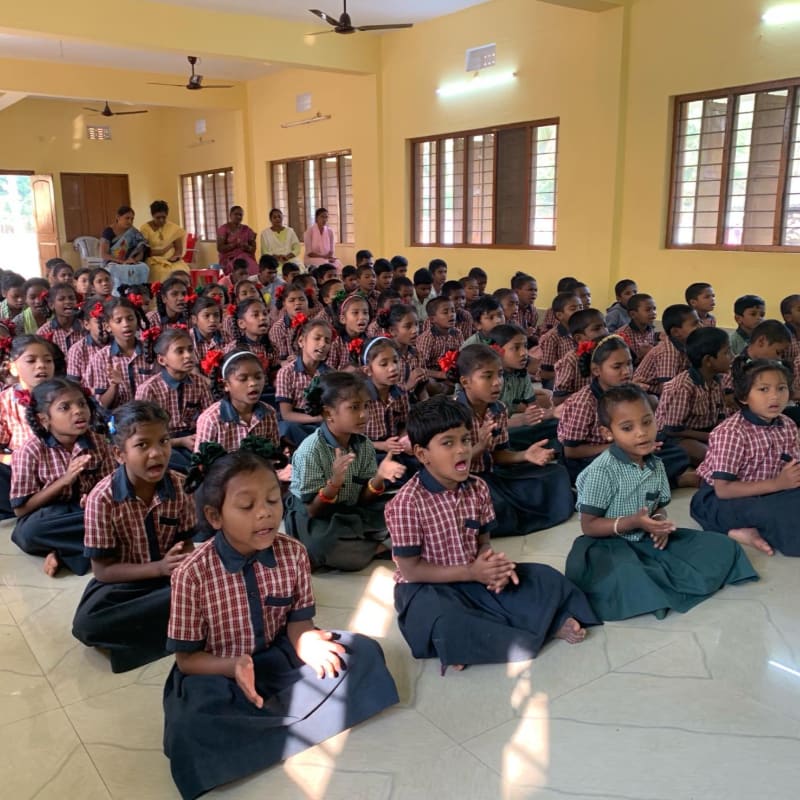 A group of school children in checkered uniforms sit on a tiled floor in the foreground, while adults sit in the background of a yellow-walled room.