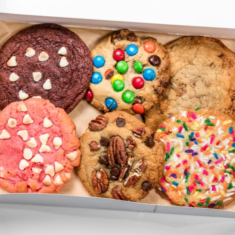 A variety of colorful cookies, including chocolate chip, M&M, and sprinkle-covered, are arranged in a box against a plain white background.