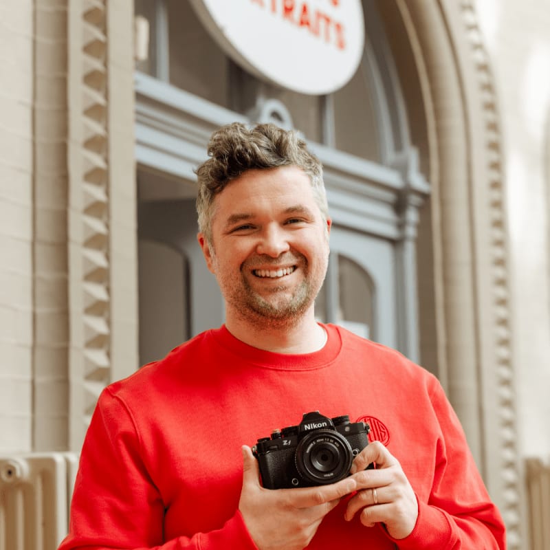 A smiling man in a red sweatshirt holds a black Nikon camera in the foreground, set against a blurred architectural background with a circular sign.