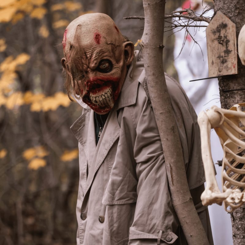 A monstrous figure in a tan trench coat stands in a wooded area next to a skeleton and a wooden marker.