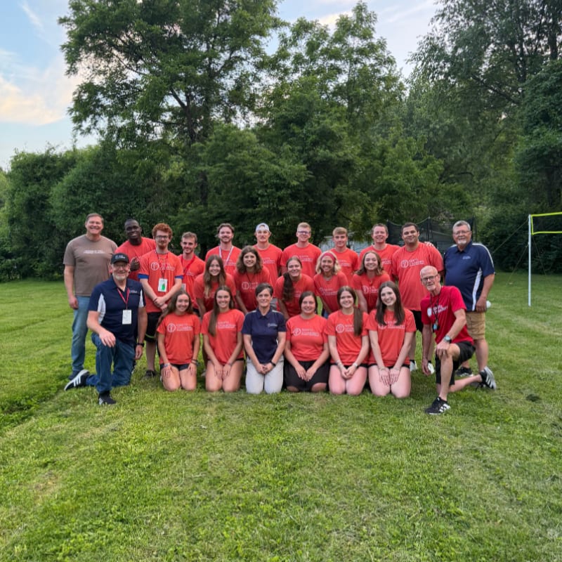 A coach in black athletic wear speaks to a group of young soccer players in red and white striped uniforms on a green field.