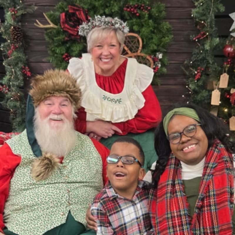 Santa Claus, Mrs. Claus, and two children smile for a photo in front of a festive holiday backdrop with greenery and ornaments.