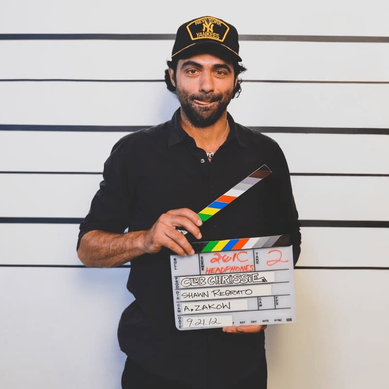 A man wearing a New York Yankees cap and a black shirt holds a movie clapperboard against a white wall with horizontal black lines.