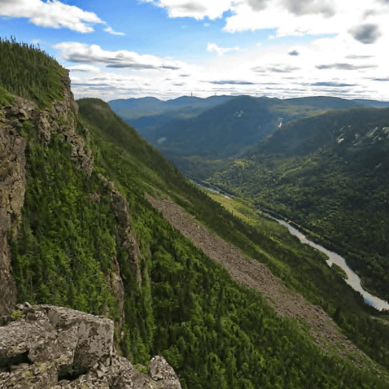 A rocky, green cliffside overlooks a deep valley with a winding river and distant blue mountains under a cloudy sky.