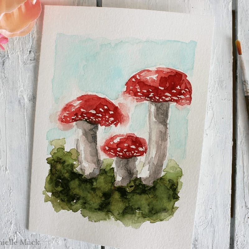 Three red-capped mushrooms with white spots grow from green foliage against a pale blue watercolor background.