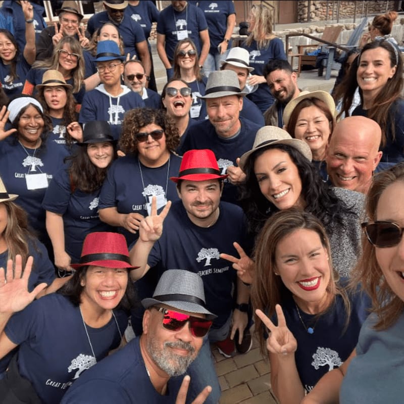 A diverse group of smiling people wearing matching navy blue t-shirts and various hats pose for a photo outdoors against a background of a building and patio furniture.