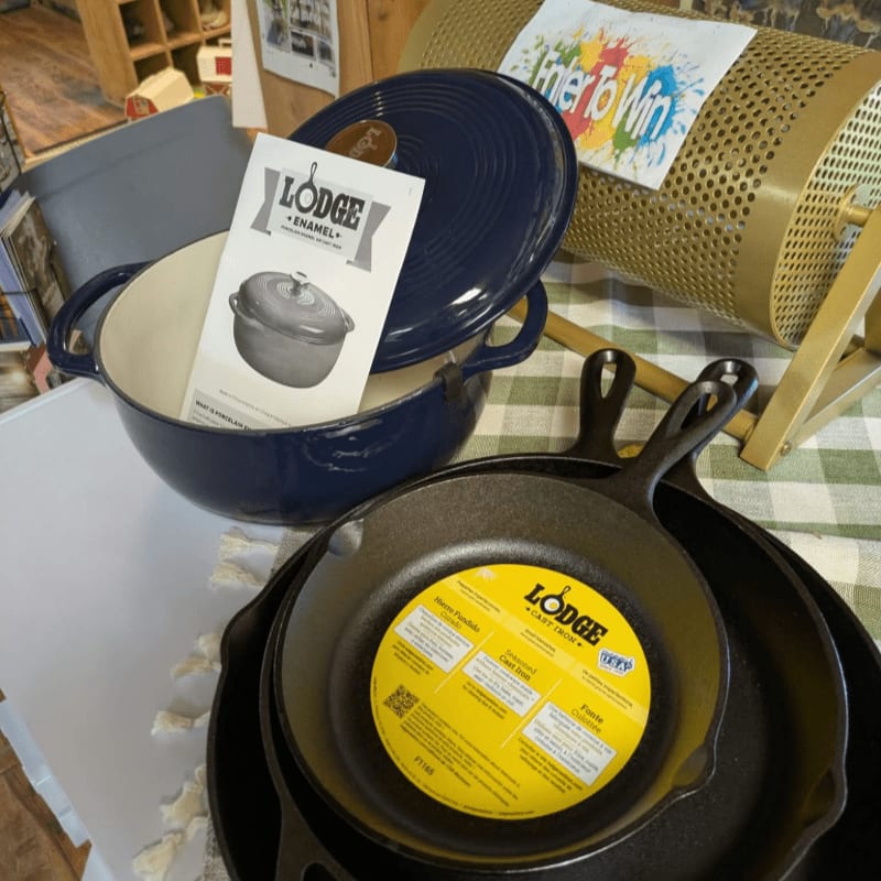A blue enamel Lodge pot and a black cast iron skillet sit in the foreground next to a gold raffle drum in the background.