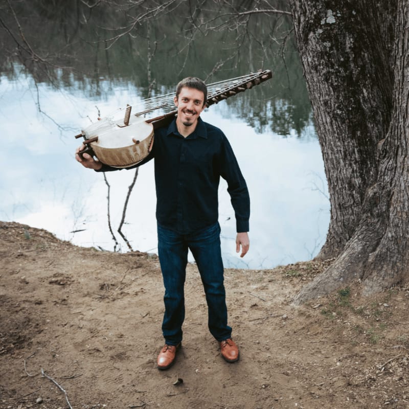 A man carrying a large stringed instrument stands on a dirt path beside a large tree and a calm body of water.