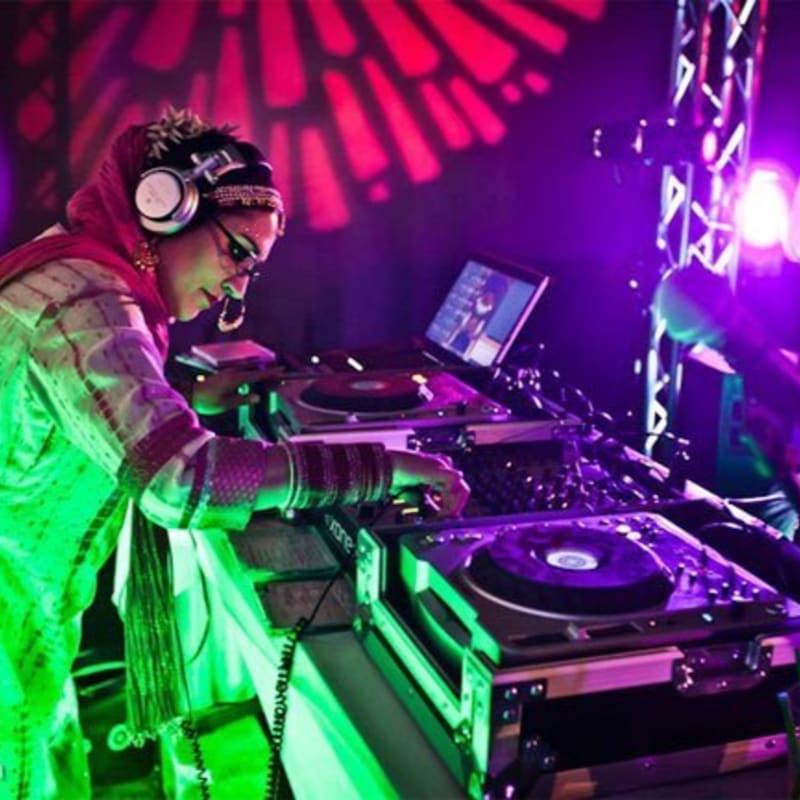 A DJ wearing traditional attire and headphones operates a mixing console in the foreground against a background of vibrant purple lighting and a red patterned projection.