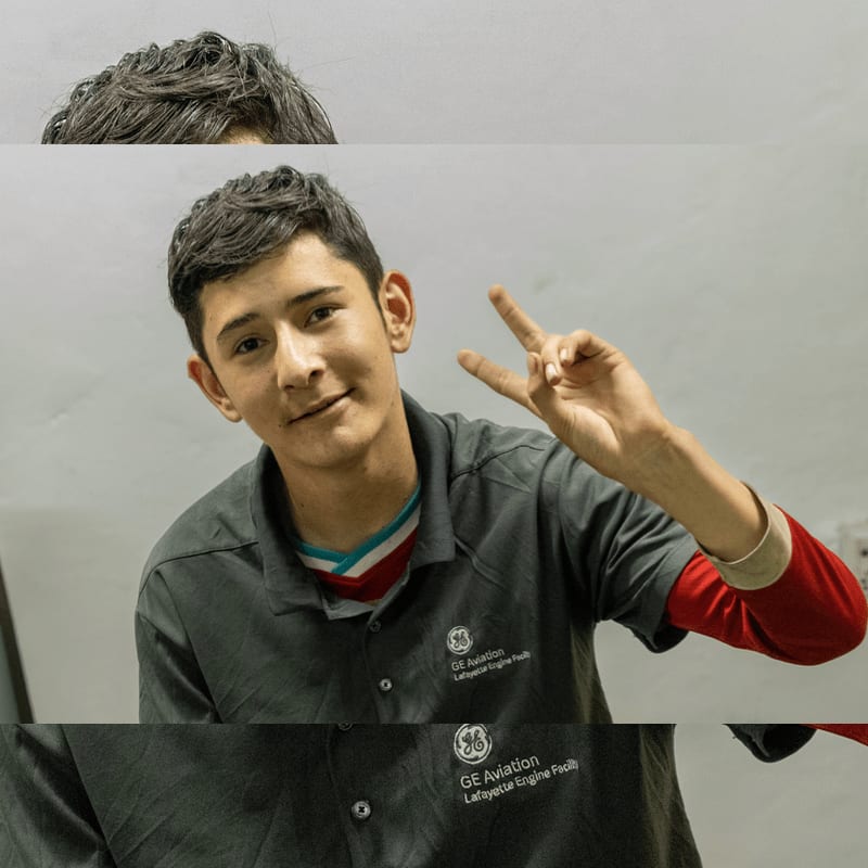 A young man in a grey GE Aviation polo shirt makes a peace sign against a plain white wall.