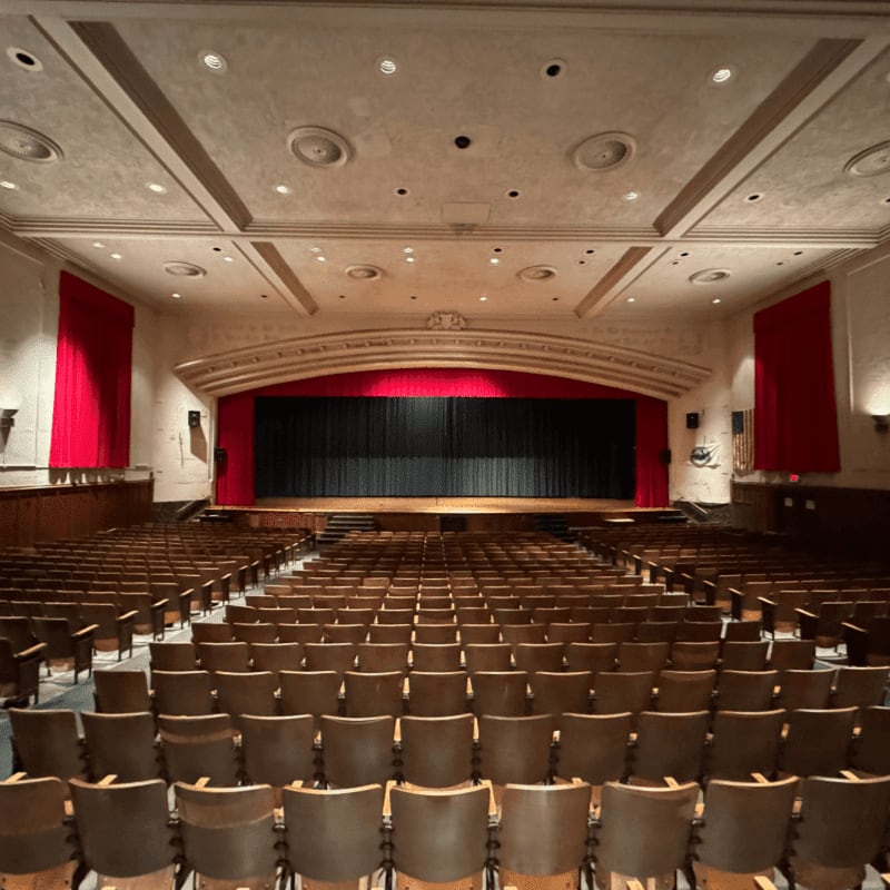 Rows of brown theater seats fill the foreground, facing a stage with a black curtain and red drapery in the background.