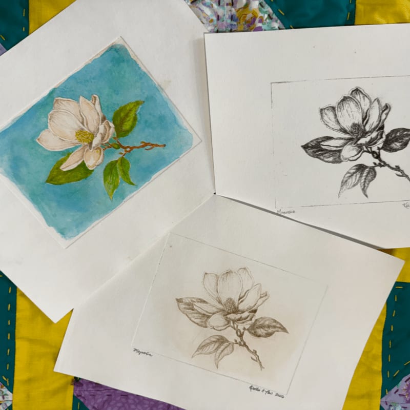 Three botanical sketches of magnolia flowers lie atop a yellow and purple quilted fabric.