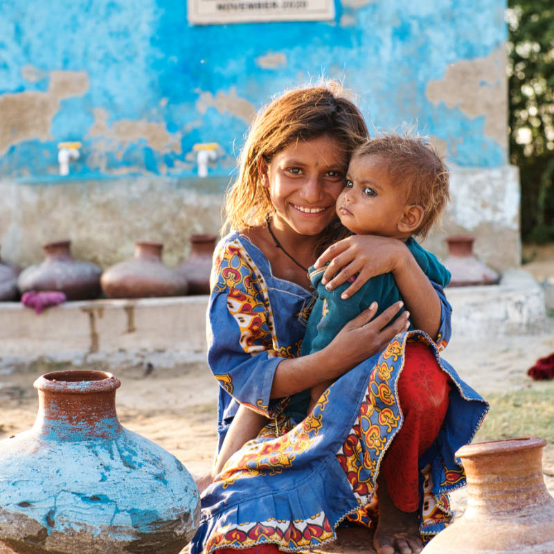 A smiling woman in a blue patterned dress holds a young child in her arms, surrounded by clay pots against a weathered blue wall.