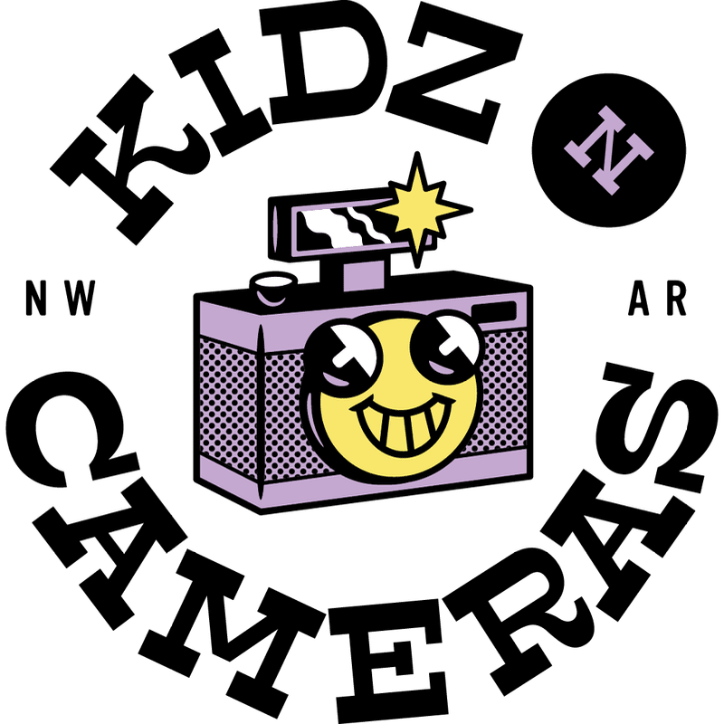 A stylized purple camera with a yellow smiley face is centered against a white background, encircled by the bold black text "KIDZ N CAMERAS."