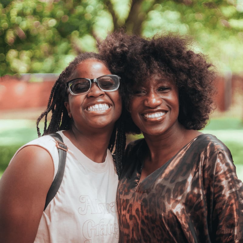 Two smiling women pose closely together in the foreground against a blurred background of green trees.