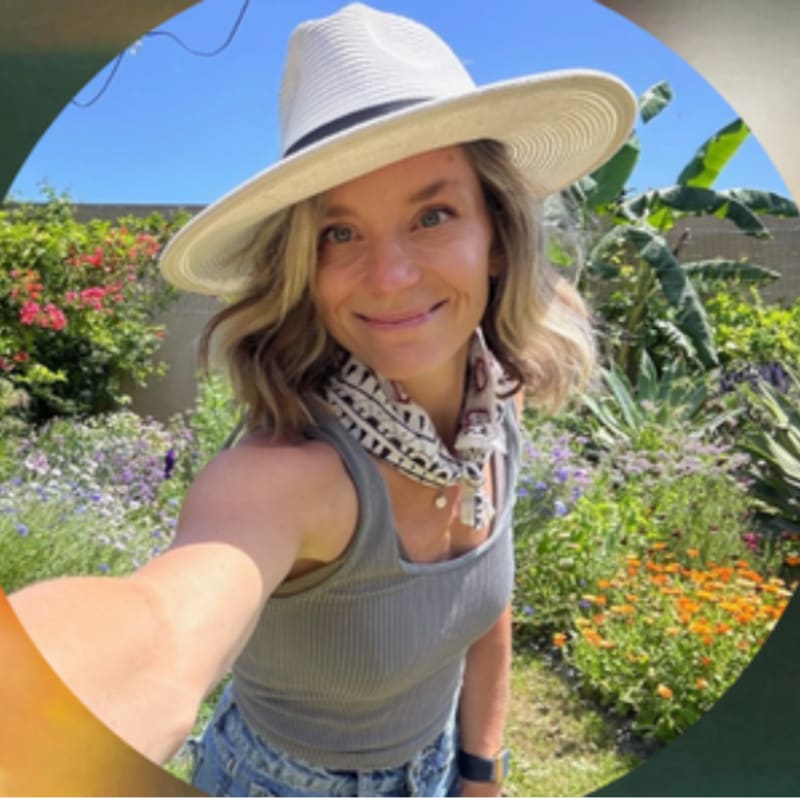 A woman wearing a wide-brimmed hat and a grey tank top smiles for a selfie in a lush garden filled with colorful flowers and green foliage.